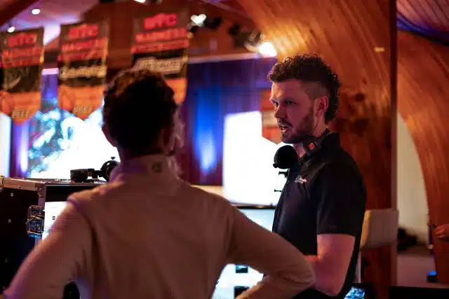 Mason Geiger talking to a woman during an event production with monitors and lights in the background.