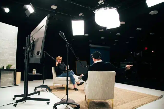 Two people sitting on a stage wearing dark clothes in tan chairs during podcast production at DreamOn Studios in Warsaw, IN.