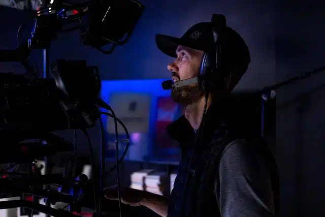 Man wearing a baseball cap with a headset on looking at a monitor during a live stream.